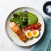 Prato low carb com salmão grelhado, ovos, abacate, tomates e folhas verdes sobre mesa de mármore para o guia de iniciantes.