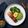 Prato com salmão grelhado, ovo, abacate e brócolis sobre mesa escura ao lado de castanhas e água com limão da marca Hábitos e Dietas.